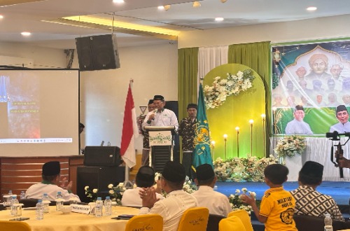 Seorang pembicara berdiri di podium dalam acara Konfercab PCNU Kotabaru 2025, dikelilingi oleh peserta dan dekorasi bertema keislaman.