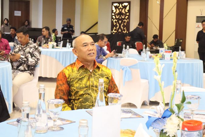Bupati Kotabaru H. Muhammad Rusli, S.Sos menghadiri acara resmi, duduk di meja dengan dekorasi biru muda, lengkap dengan botol air mineral dan hiasan bunga.